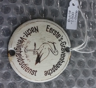 0000-0003-MMO.jpg; 0000-0003-MMO; Geëmaillerd rond naamplaatje met Haagse ooievaar en de tekst "Eerste 's-Gravenhaagsche Nacht-Veiligheidsdienst".; naamplaten